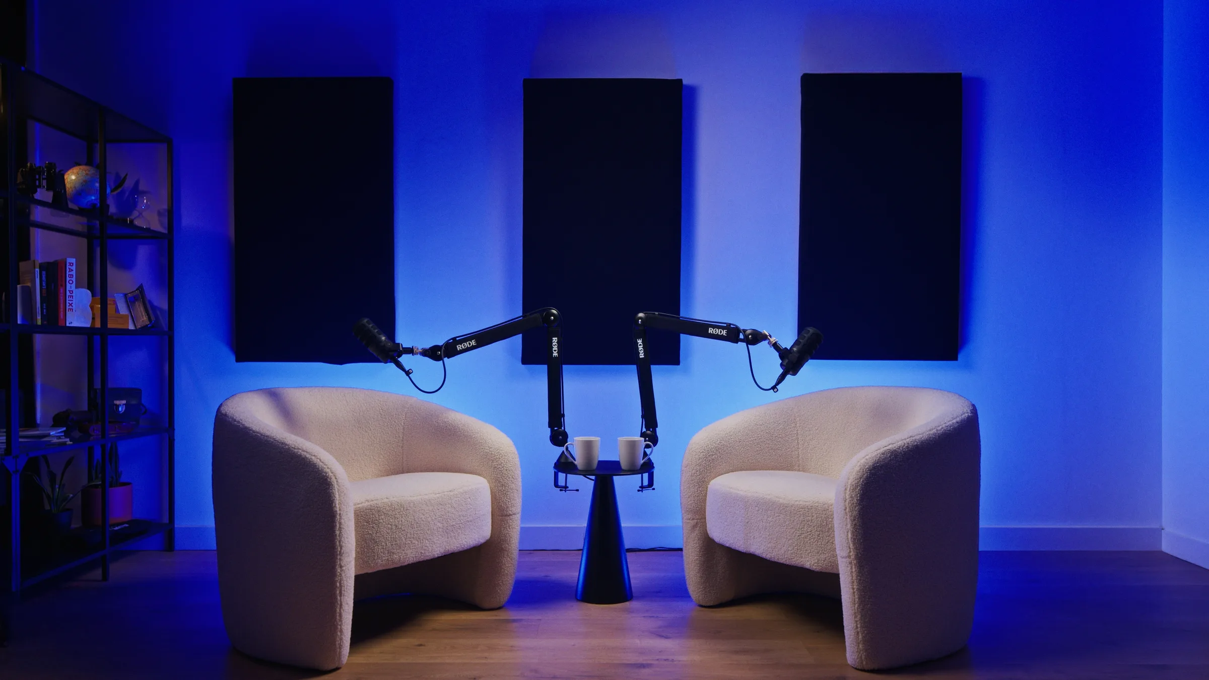 Setup de podcast no estúdio AIRLUSO: duas poltronas brancas, mesa de apoio, microfones RØDE e painéis acústicos na parede com iluminação azul ambiente.
