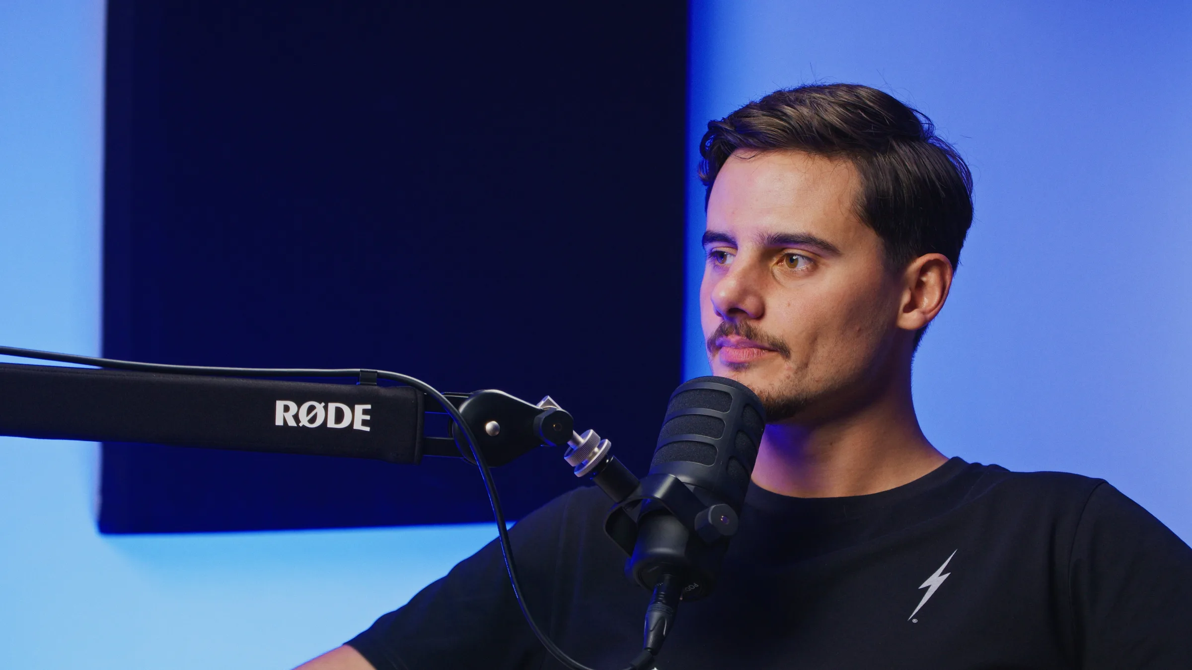 Convidado em podcast — t-shirt preta, microfone RØDE em close-up, iluminação azul de fundo.